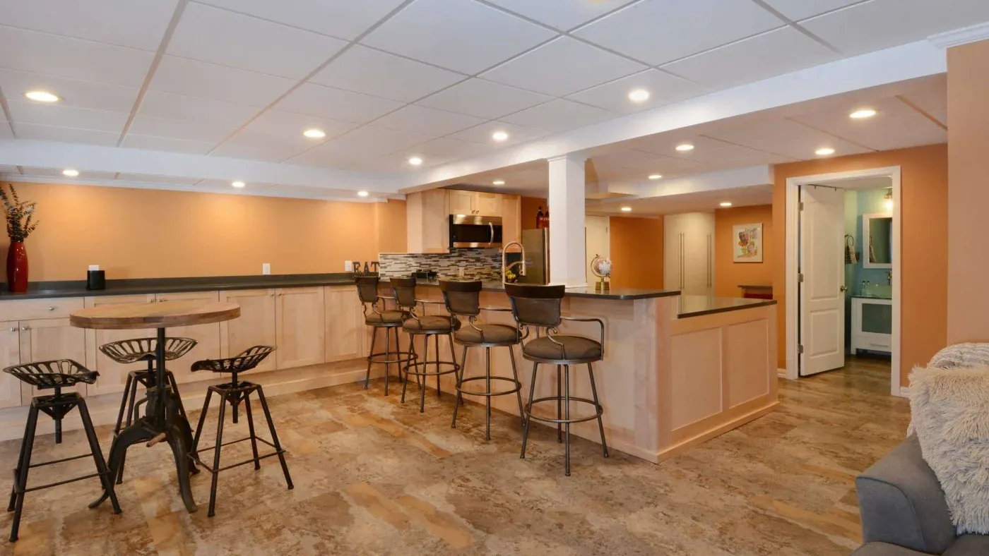 Spacious kitchen area with warm beige tones and tiled flooring. Features a bar counter with metal stools, a round table with metal chairs, and modern lighting. Cozy and inviting atmosphere.