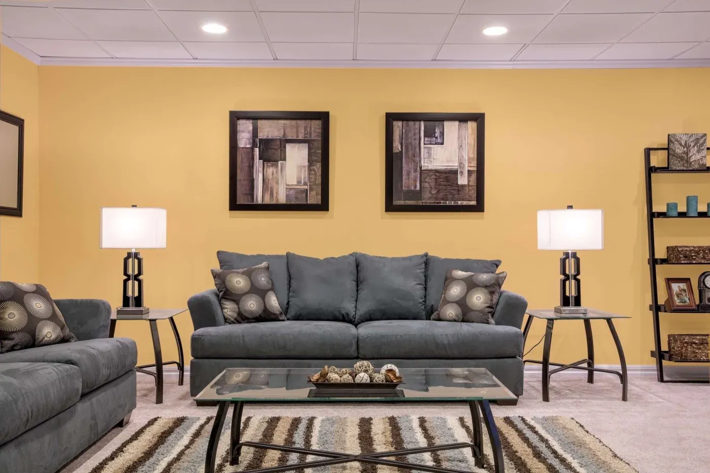 A cozy living room with a gray sofa, patterned cushions, and matching armchairs against a yellow wall. Two table lamps and abstract art add warmth.