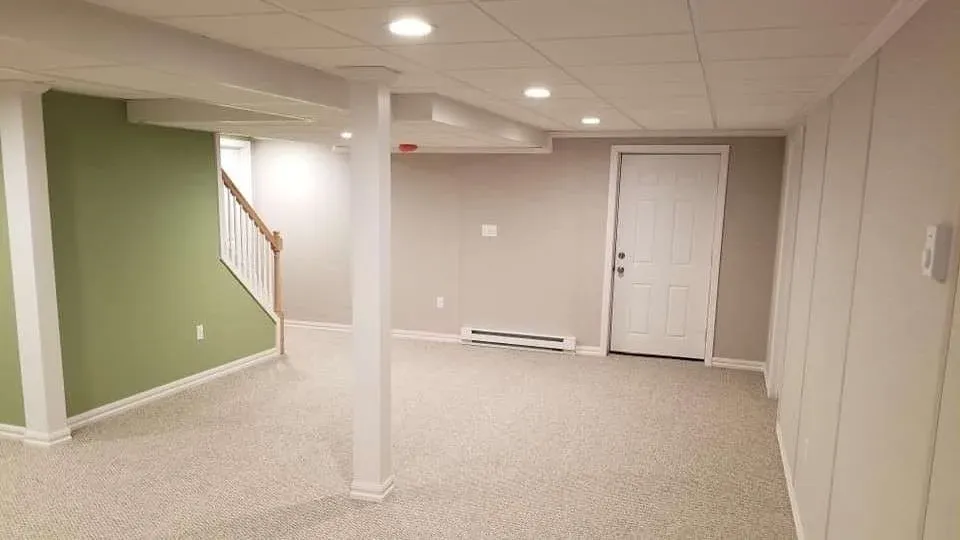 A spacious basement with beige carpet, pale green accent wall, white pillars, recessed lighting, and a staircase. The atmosphere is clean and modern.