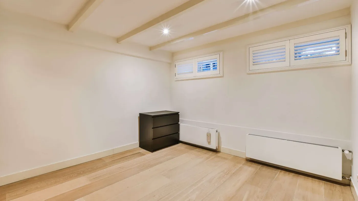 A minimalist room with light wooden flooring, two small windows with blinds, and a black dresser. The space has neutral walls and a calm, spacious feel.