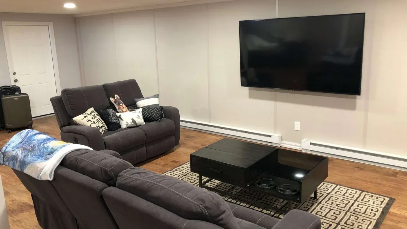 A cozy living room with gray sofas, patterned cushions, and a throw blanket. A large flat-screen TV is mounted on the wall. A black coffee table sits on a geometric rug, creating a modern and inviting atmosphere.