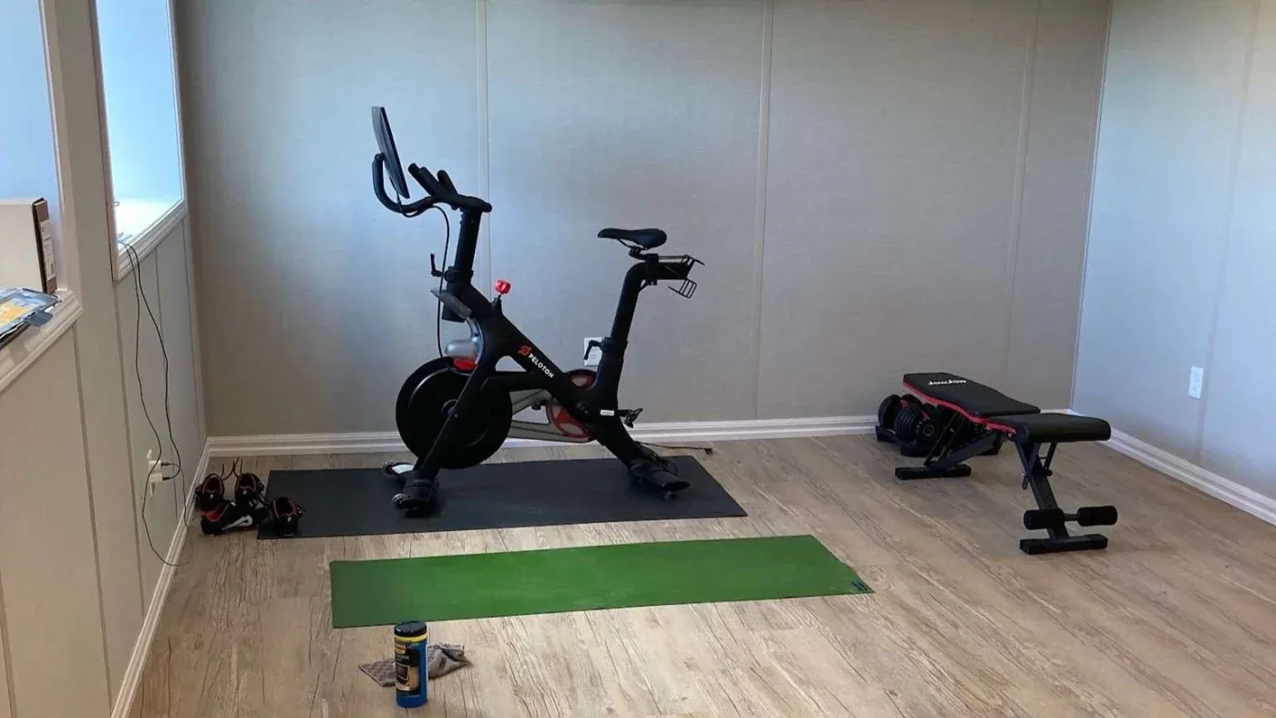 Compact home gym with a stationary bike on a mat, a green yoga mat, and an adjustable bench with dumbbells nearby. Bright, organized, and functional space.