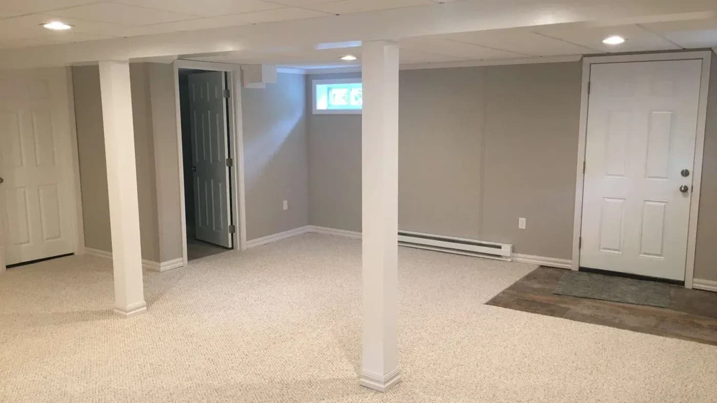 A clean, empty basement with beige carpet, light gray walls, and white ceiling. Two white doors and small windows add brightness. Calm, neutral feel.
