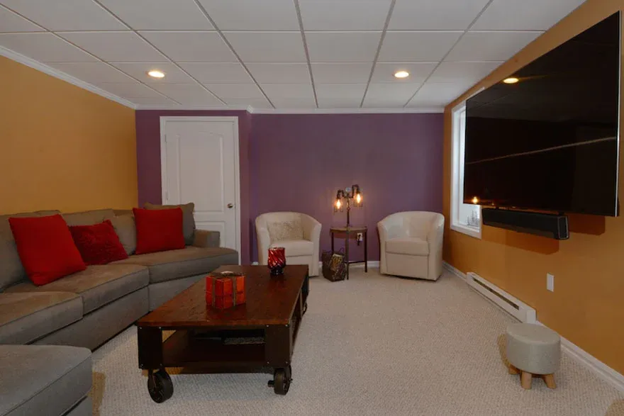 A cozy basement living room with a large gray sectional sofa adorned with red cushions. There's a wooden table with candles, two white chairs by a purple accent wall, a modern floor lamp, and a big flat-screen TV mounted on a mustard-colored wall. The space feels warm and inviting.