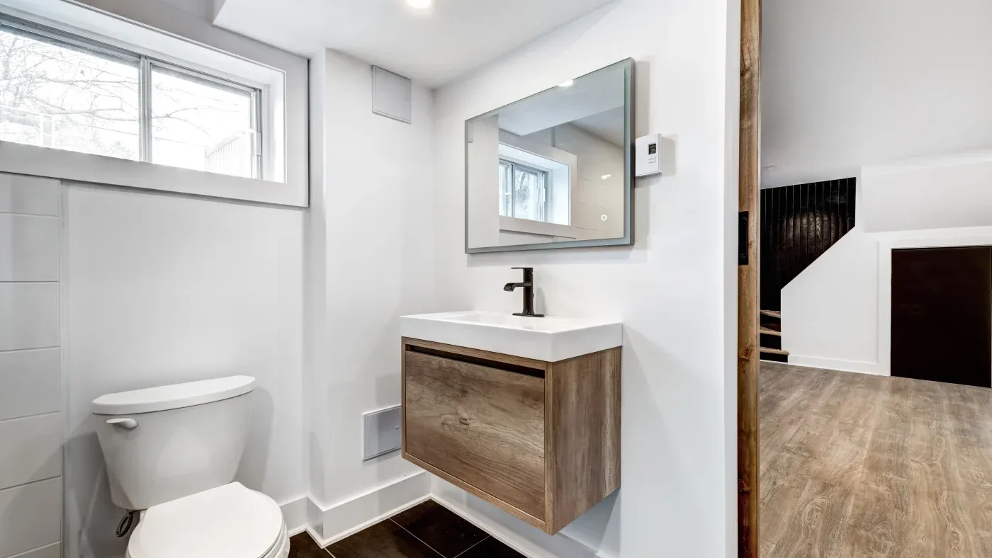 Modern bathroom with a wall-mounted sink featuring wooden cabinetry, a sleek faucet, and a large mirror. Adjacent is a white toilet and a window.