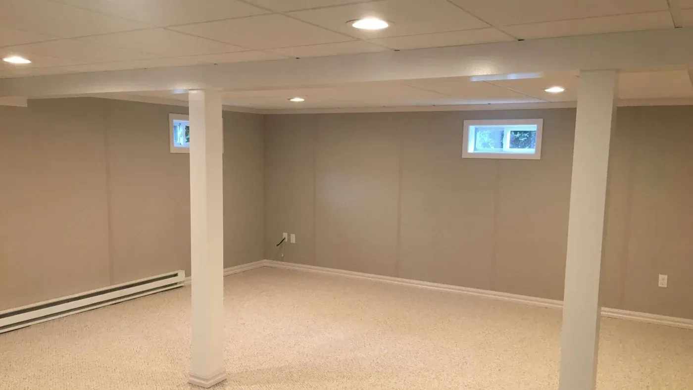 A spacious, empty basement with beige walls and carpeted flooring. Two small windows let in natural light. The room has recessed ceiling lights and white support beams.