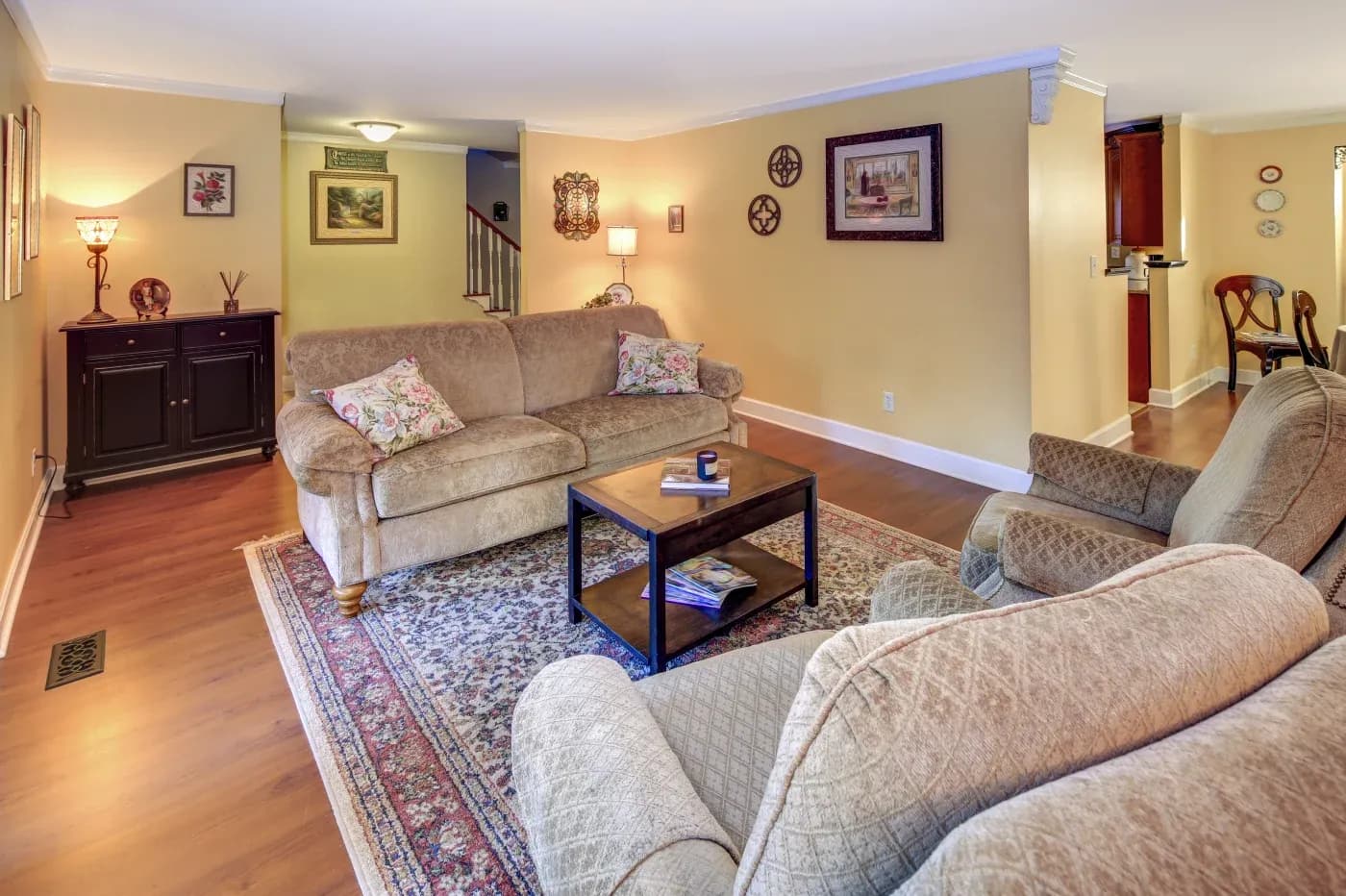 A cozy living room with beige couches, patterned cushions, and a wooden coffee table on a decorative rug. Warm lighting and wall art create an inviting atmosphere.