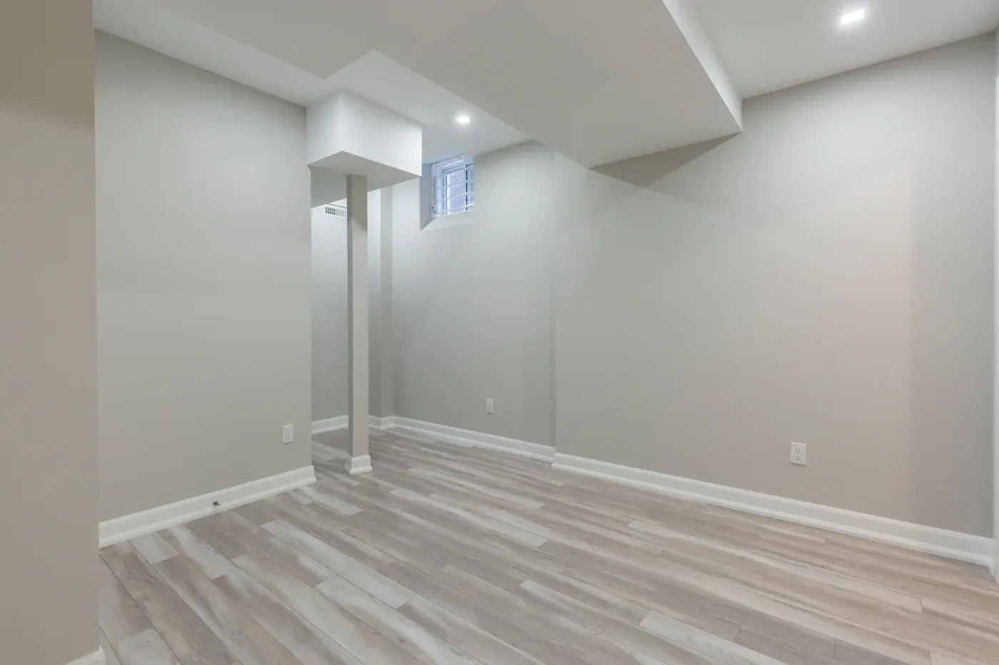 A modern, small, empty room with light gray walls and wood flooring. The ceiling has recessed lights and a small window, giving a clean, minimalist feel.