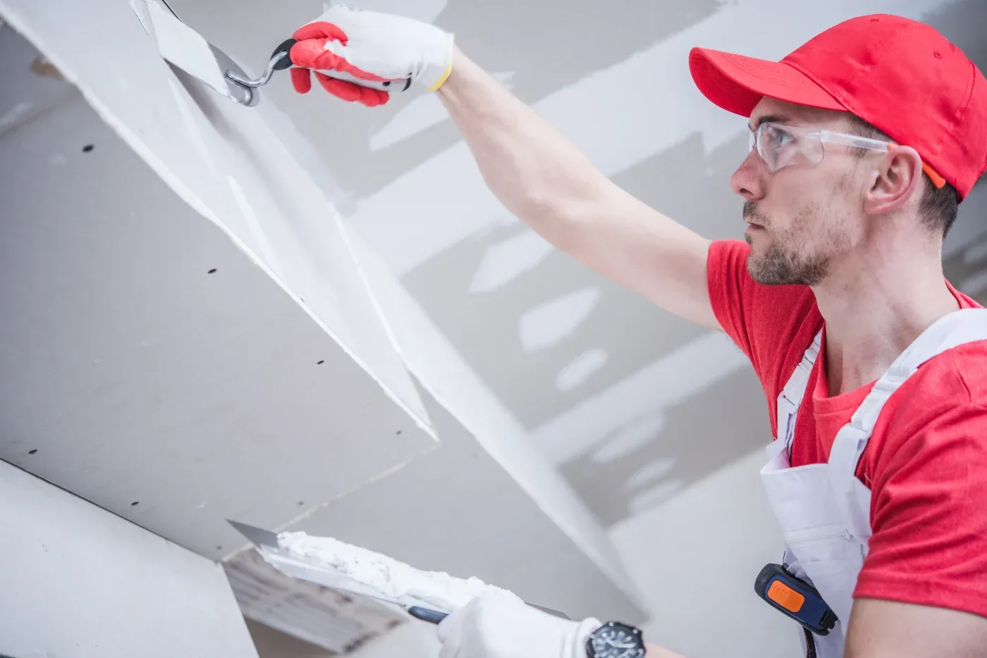 A focused worker in a red cap and glasses applies plaster to a ceiling with a trowel, wearing white overalls and gloves, conveying concentration.