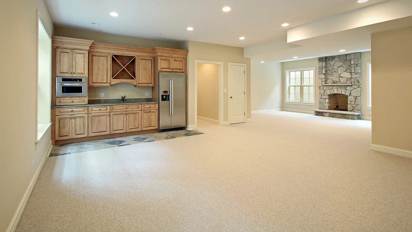 Spacious modern basement with beige walls and carpet. Features a kitchenette with wooden cabinets, stainless steel appliances, and a stone fireplace.
