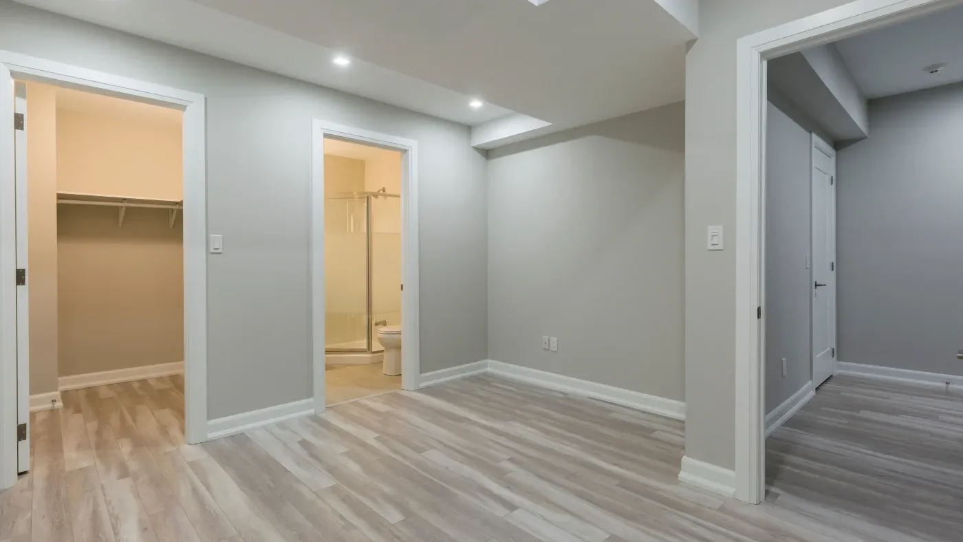 Spacious, empty room with light gray walls and laminate flooring. Doorways lead to a closet and bathroom with shower. Bright, modern, and minimalistic.
