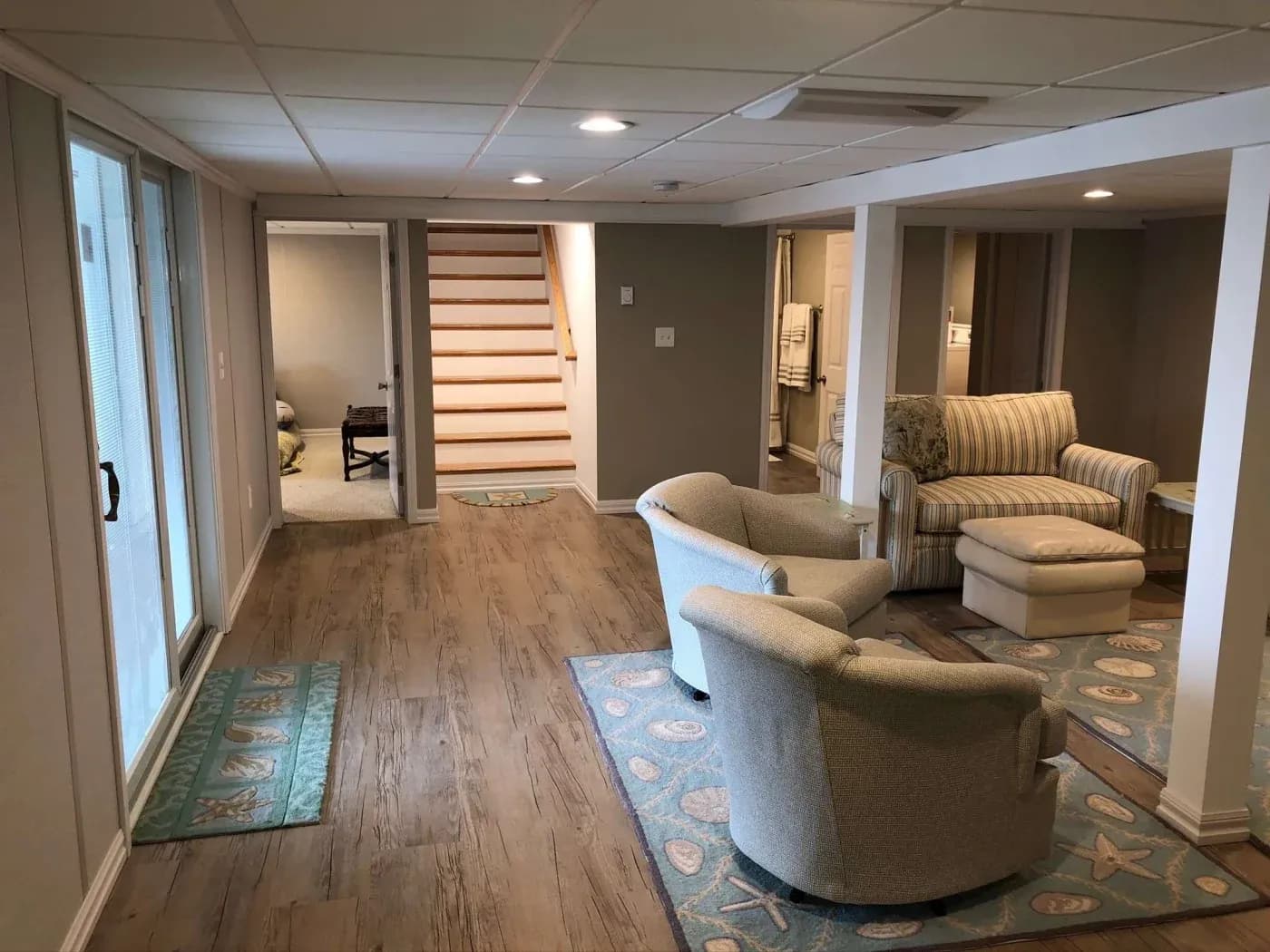 Basement family room with wood floors, beige walls, and recessed lighting. Two beige armchairs and a striped couch on patterned rugs create a cozy feel.