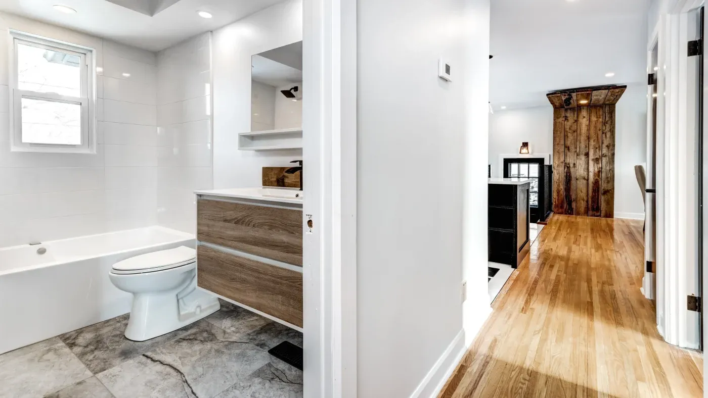 Modern bathroom and hallway; bathroom features a sleek design with marble floor, white fixtures, wooden vanity, and a bright window. Hallway has warm wooden floors, white walls, and rustic wooden accents, conveying a cozy yet contemporary feel.