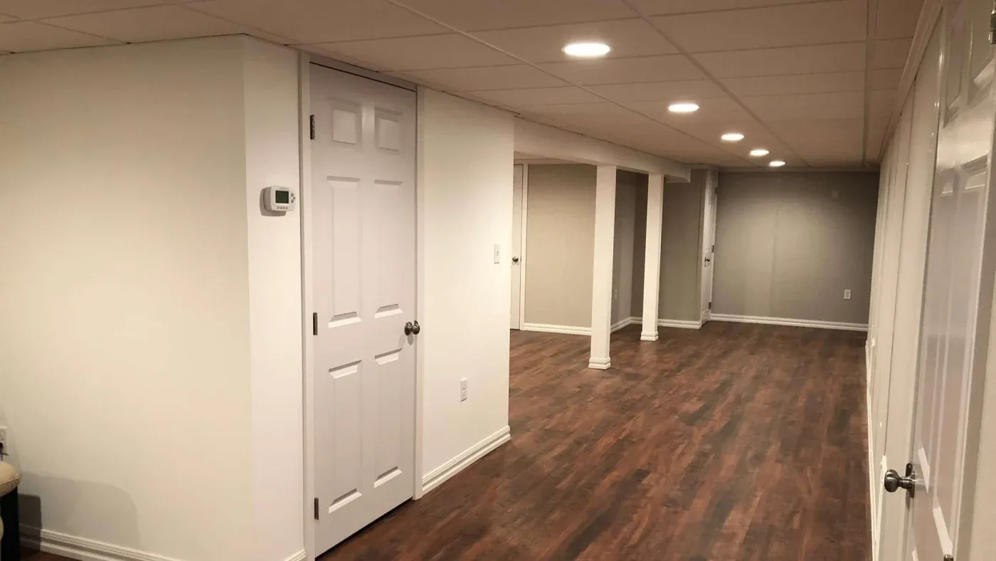 Spacious basement with white walls and wood flooring, featuring recessed ceiling lights. A row of support columns aligns the central area.