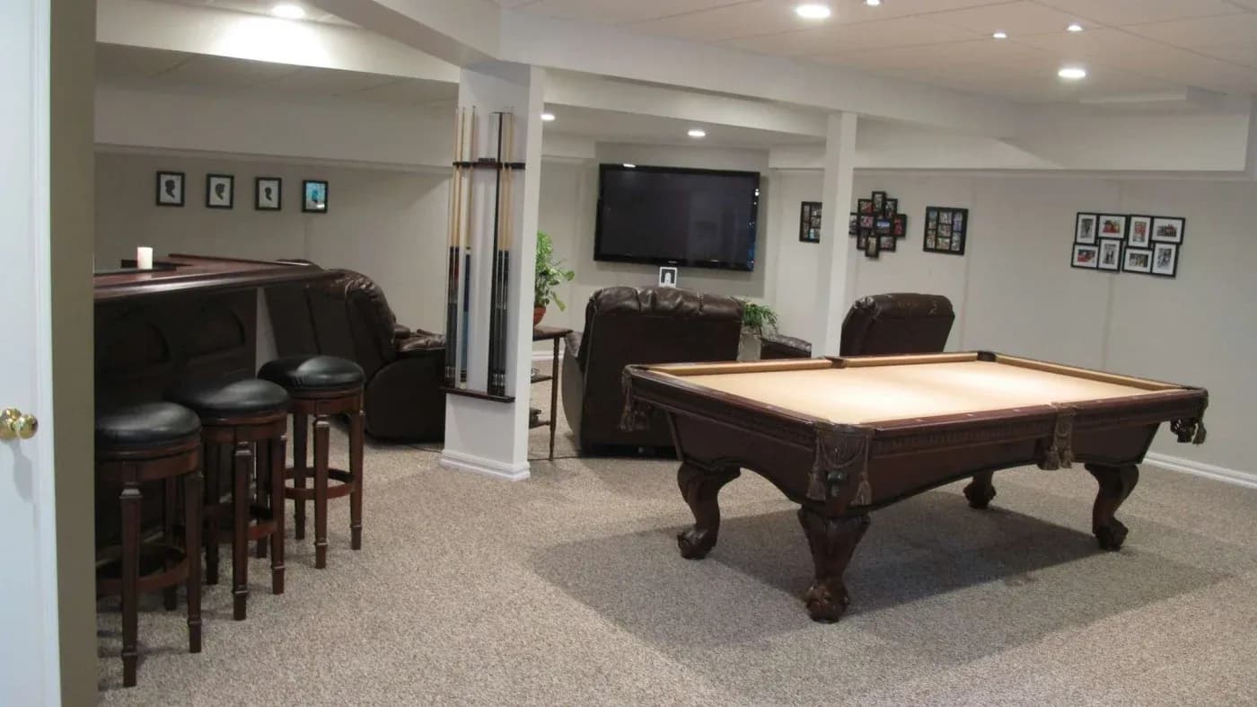 A cozy basement game room features a billiard table, leather recliners facing a large TV, and a bar area with stools. The walls are adorned with framed photos.
