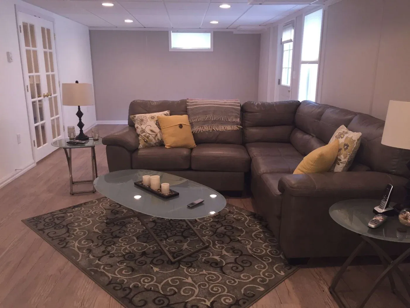 Cozy living room with a brown leather sectional sofa adorned with yellow and patterned cushions, beside glass tables, and decorative candles. Warm, inviting ambiance.