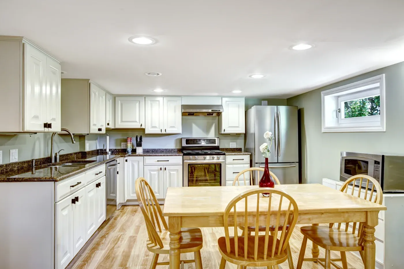 Bright kitchen with white cabinets, stainless steel appliances, and granite countertops. A wooden table with four chairs centers the room, under recessed lighting.