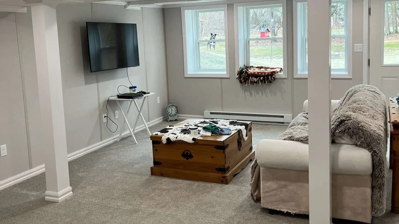 A cozy basement room with a wall-mounted TV, a small table, and a cowhide rug on a wooden trunk. Large windows let in natural light. Neutral tones create a calm atmosphere.