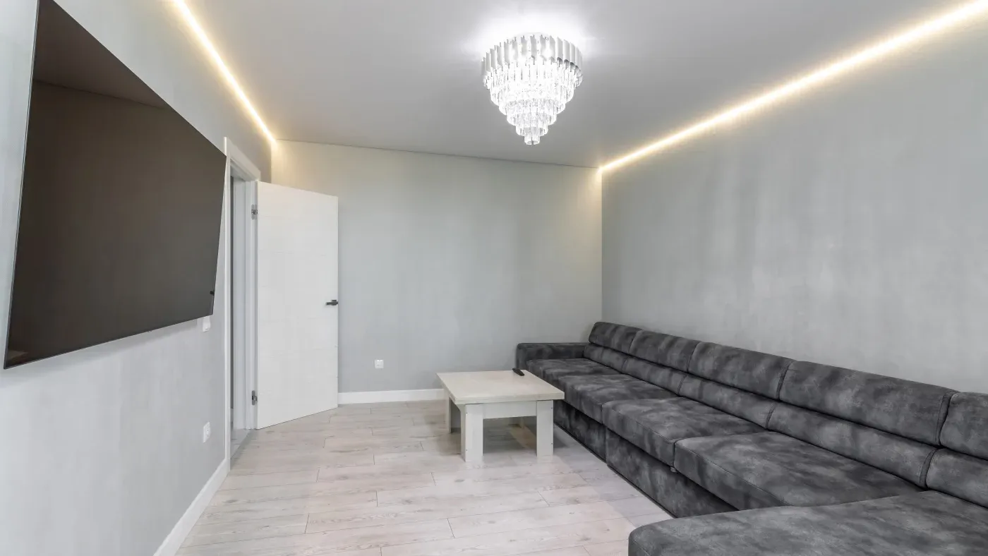 Modern living room with gray walls, a large L-shaped gray couch, a white door slightly open, a sleek coffee table, and a sparkling chandelier.