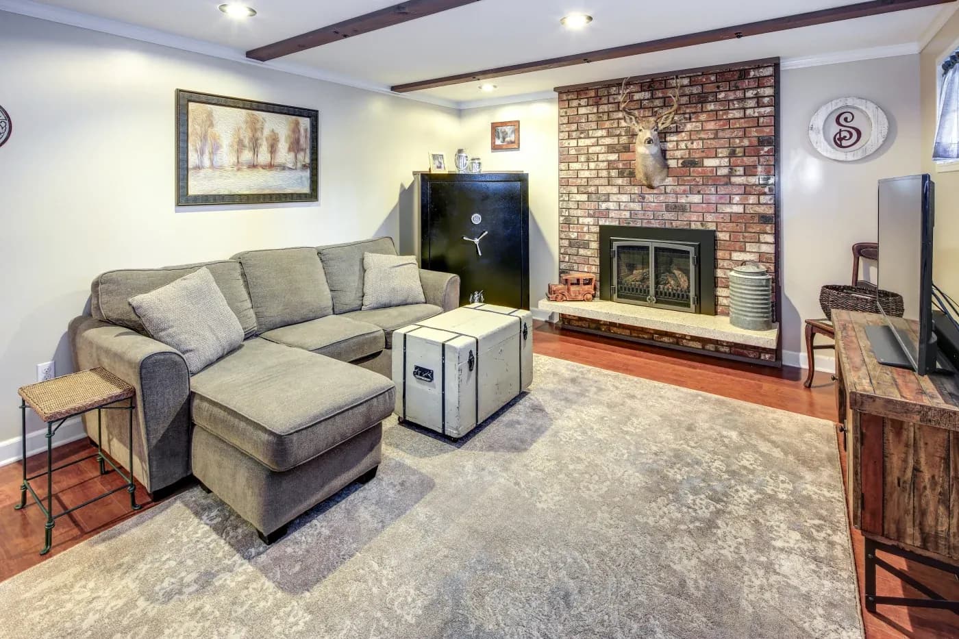 Cozy living room with a sectional sofa, vintage trunk as a table, brick fireplace, and deer head mount. Soft, warm lighting creates a welcoming ambiance.