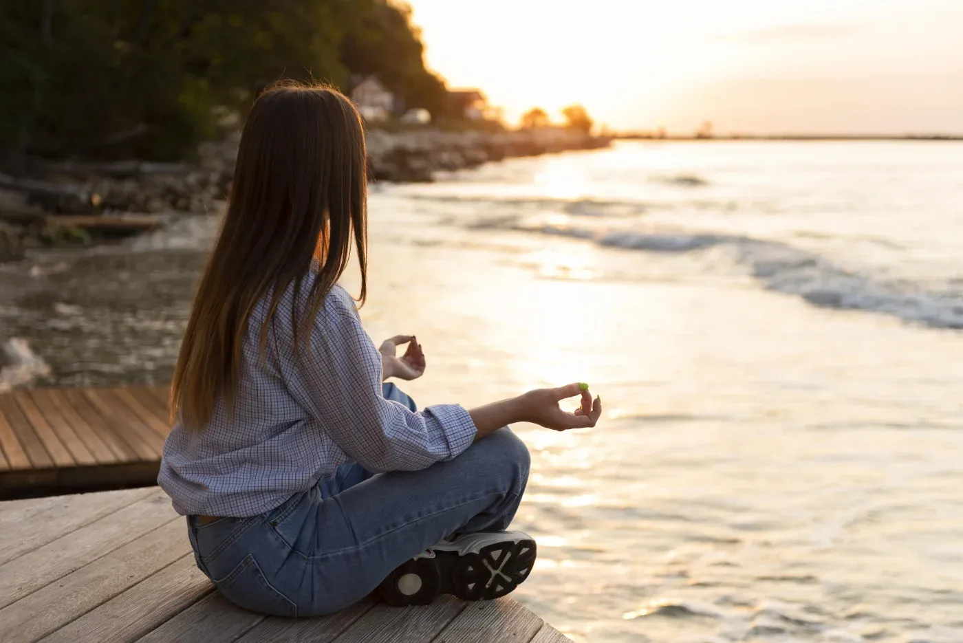 A woman sits cross-legged on a wooden dock, meditating by the water at sunset. The scene conveys tranquility with orange hues reflecting on the waves.