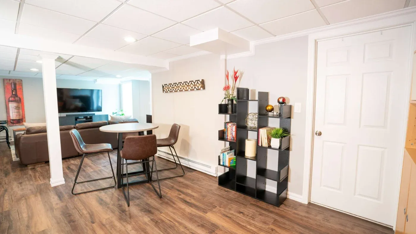 Modern basement room with wooden floors, featuring a round table and four chairs. A black cube shelf with books and plants decorates the wall, and a TV setup with a brown sofa is in the background, creating a cozy and stylish atmosphere.