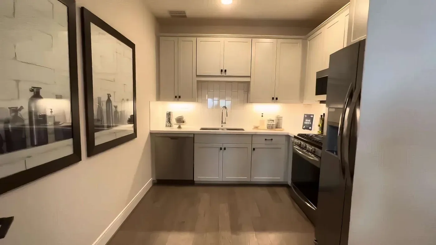 A modern, compact kitchen with white cabinets, stainless steel appliances, and wooden flooring. Two framed artworks adorn the left wall, adding elegance.
