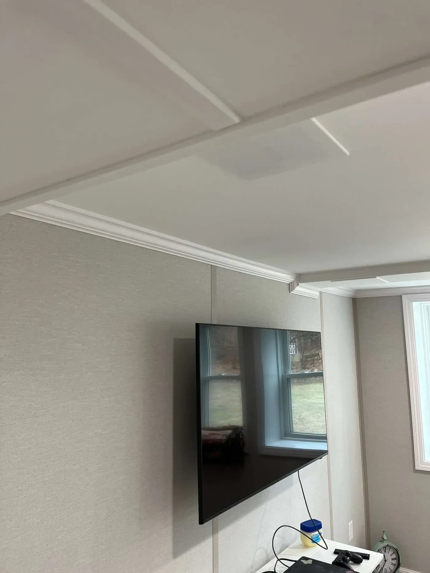 Wall-mounted TV in a modern living room with beige walls and crown molding. A window reflects the outdoors. A game controller and cup are on a nearby table.