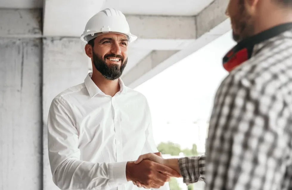 A bearded man in a white shirt and hard hat smiles while shaking hands with another man in a checked shirt, suggesting a successful meeting at a construction site.