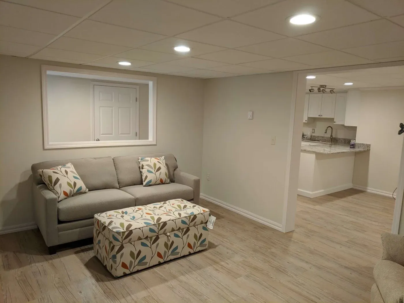 A cozy basement living area with a beige sofa adorned with floral cushions. A patterned ottoman matches the cushions. Neutral walls and modern lighting.