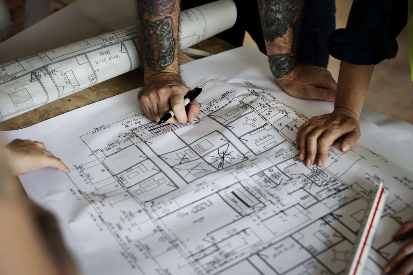 Hands of several people, some tattooed, are reviewing and marking architectural blueprints with a pen on a wooden table, conveying teamwork and focus.