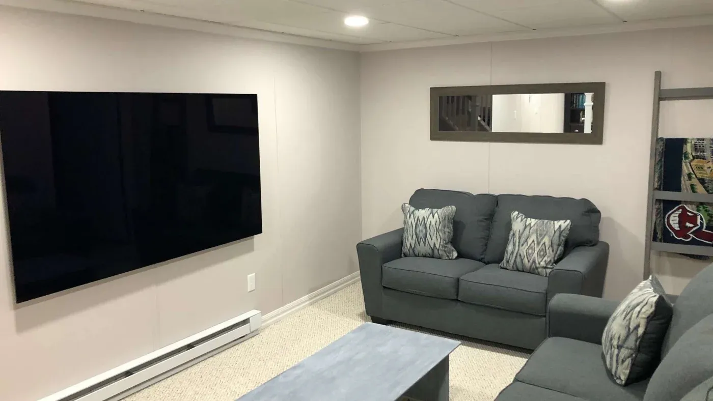 A cozy living room with gray sofas adorned with patterned cushions, a large flat-screen TV on the wall, and a ladder shelf holding books. Emphasizes comfort.