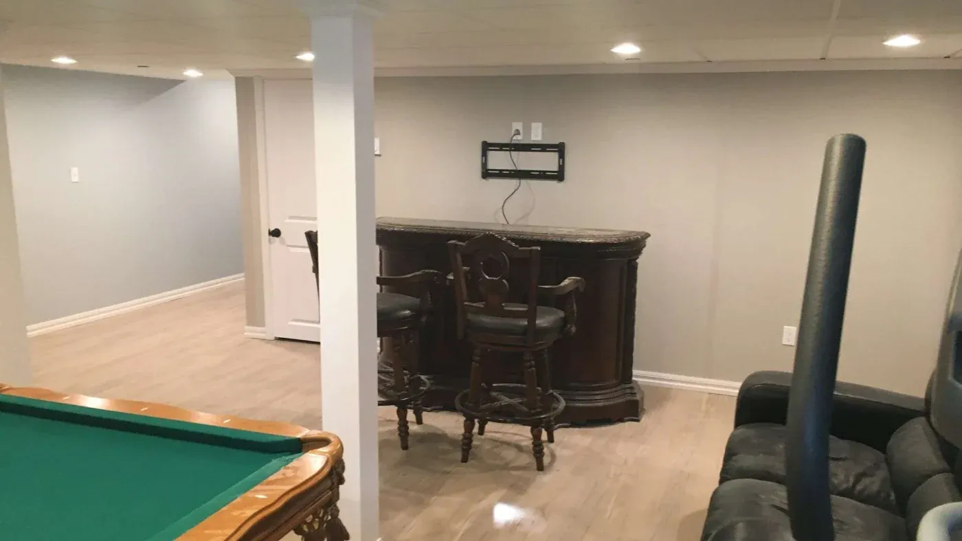 A modern basement features a pool table with a green surface, a small bar with two chairs, a wall mount for a TV, light wood flooring, and cozy seating.