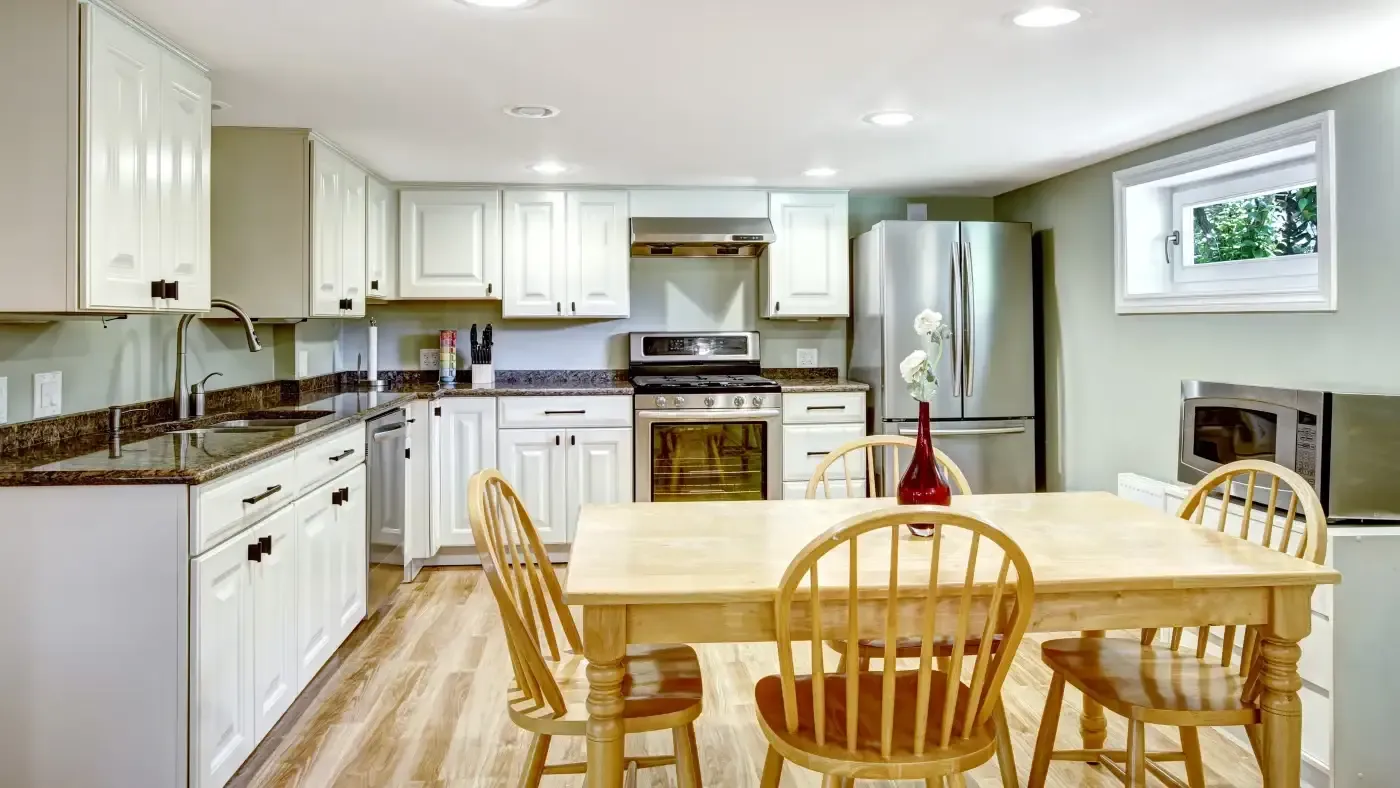 A cozy kitchen with white cabinets, stainless steel appliances, and wooden floor. A light wood dining table with a red vase and four chairs sits centrally.