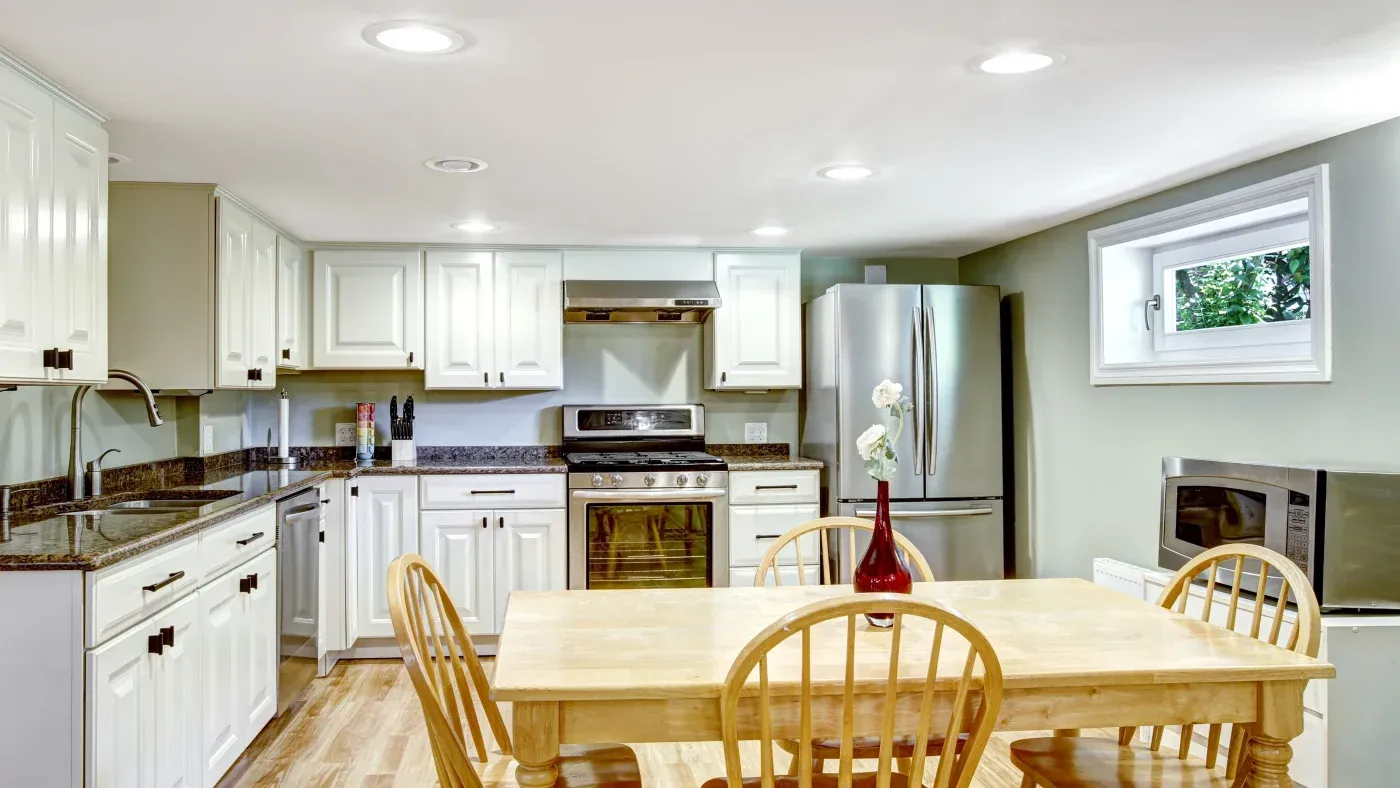 A bright kitchen with white cabinets, stainless steel appliances, and a wooden dining table. A window adds natural light, creating a welcoming atmosphere.