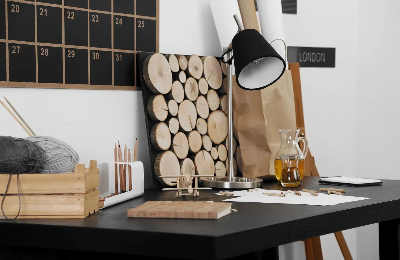 Modern workspace with a black desk featuring a table lamp, wooden art piece, and a calendar. Art supplies and a glass jug add a creative touch.