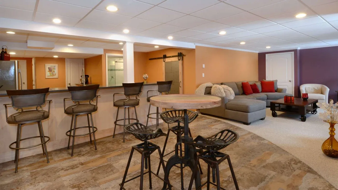 Spacious basement with a modern industrial style. Features a curved wooden bar with metal stools, a gray sectional sofa with red cushions, and warm lighting.