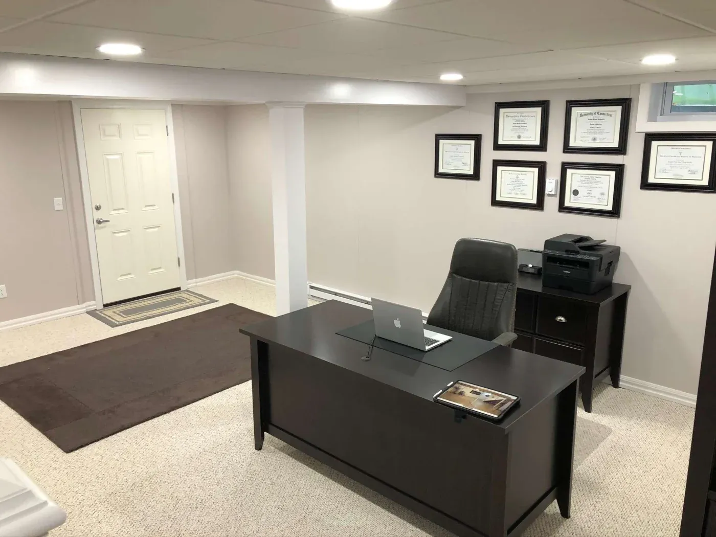 A well-organized office with a dark desk, laptop, brown chair, and a printer on a side table. Certificates on the wall suggest professionalism.