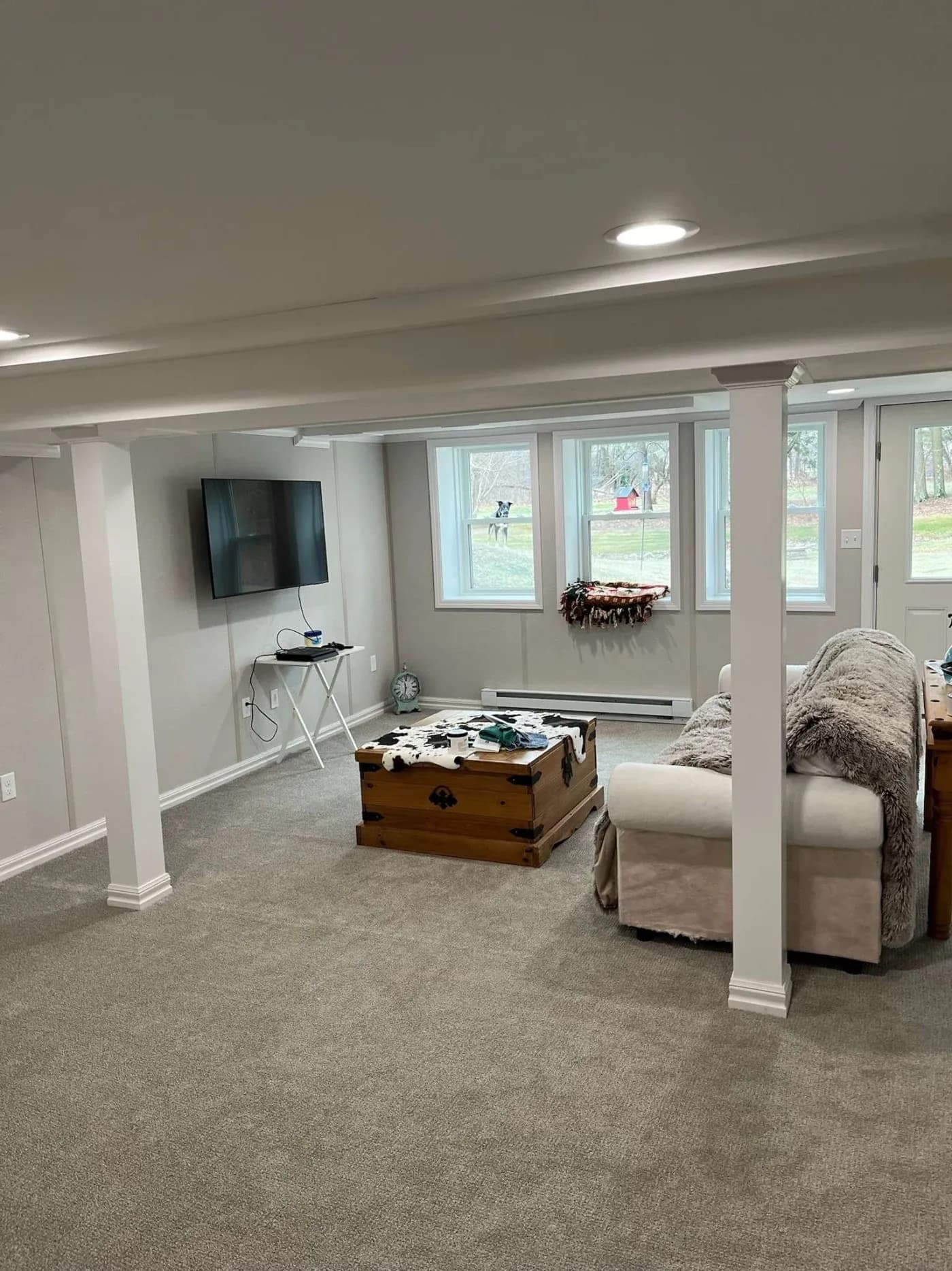 A cozy basement with gray carpet and white walls features a sofa with plush pillows, a wooden chest, a wall-mounted TV, and large windows.