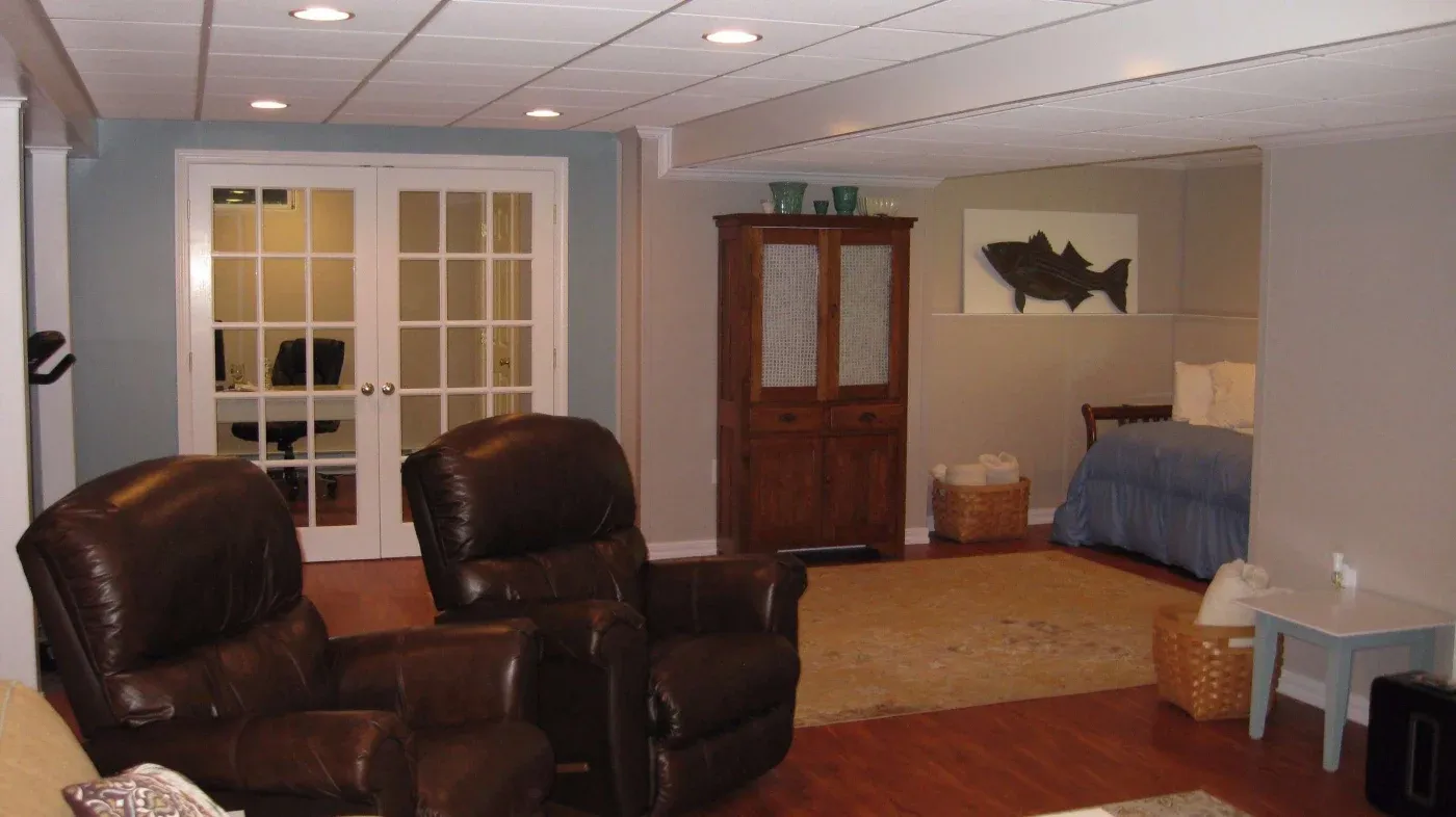 A cozy basement living area with brown leather recliners on wooden floors. Nearby, a small bedroom with a blue bedspread and decorative cabinet.