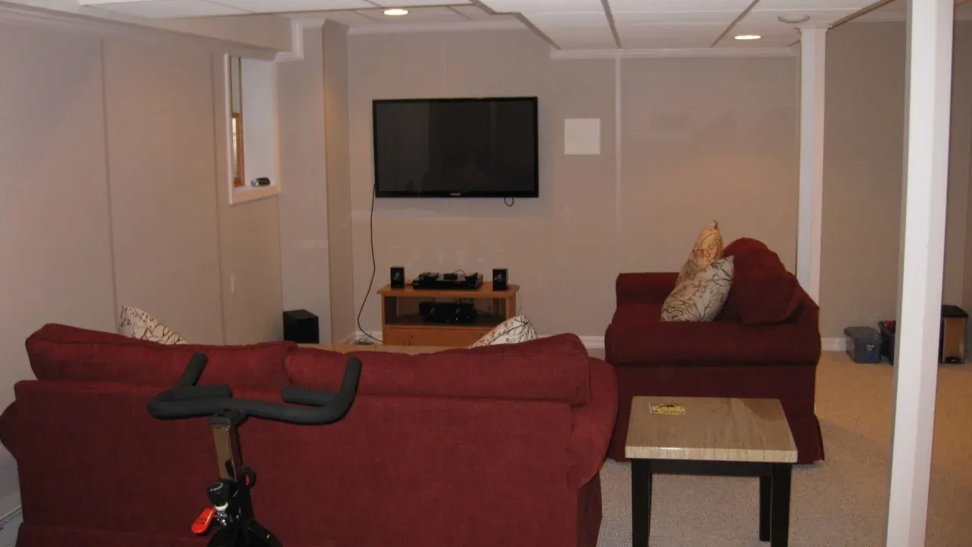 Basement room with maroon sofas facing a wall-mounted TV, beside a small wooden table. An exercise bike is partially visible, creating a cozy atmosphere.