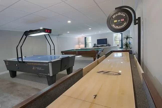 Recreation room with air hockey, shuffleboard, pool table, and TV. Brightly lit, relaxed atmosphere for games and entertainment.