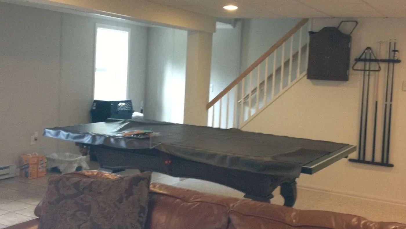 A basement room with a covered pool table, leather sofa, and a staircase. Pool cues and a rack are mounted on the wall, creating a cozy leisure space.