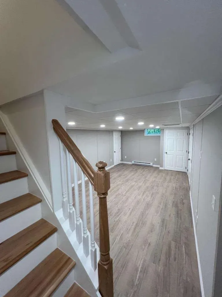 A newly renovated basement with wooden floors and white walls, featuring recessed lighting. A staircase with wooden handrails is visible on the left.