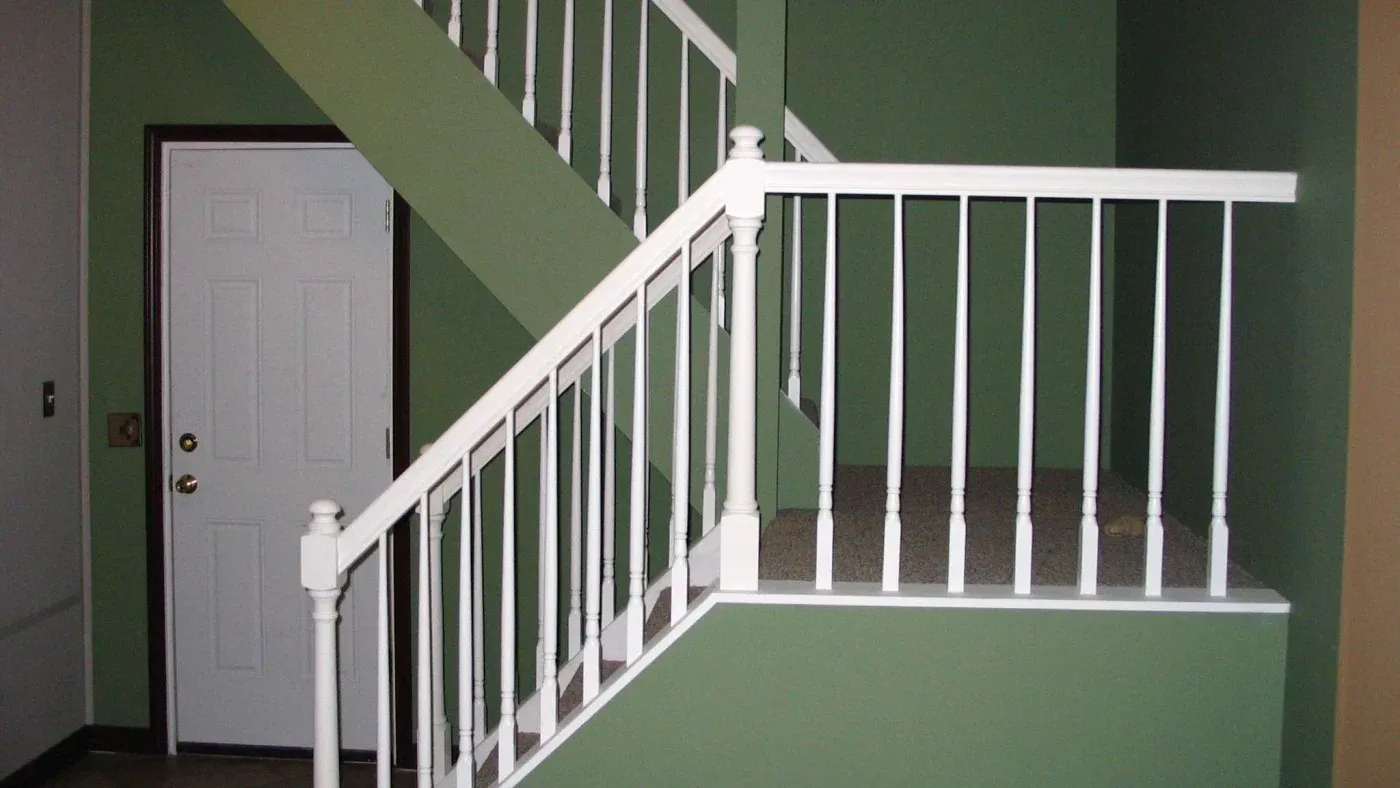 A white railing staircase set against olive green walls. A white door with a brass handle is seen to the left, creating a clean, minimalistic look.
