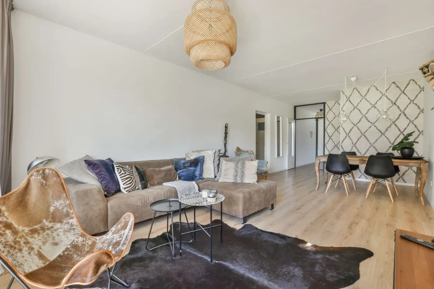 A stylish living room with a beige sectional sofa decorated with pillows, a cowhide chair, a black coffee table, and a pendant light. A dining area with a wooden table and black chairs is visible. Light wood flooring and a patterned accent wall create a cozy, modern vibe.