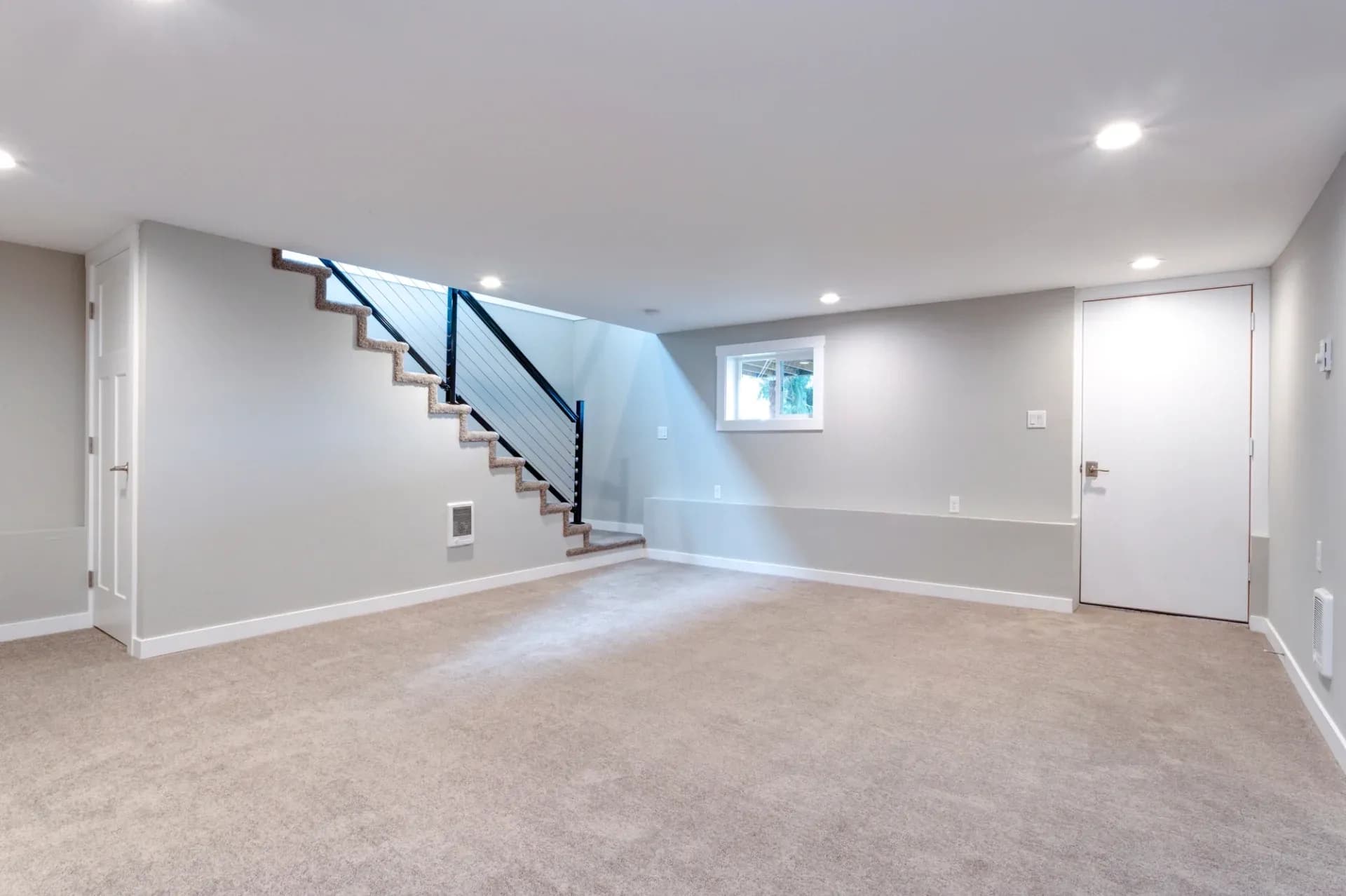 A spacious, carpeted basement with neutral light gray walls, recessed ceiling lights, and a staircase with dark railings. A door and small window are visible.