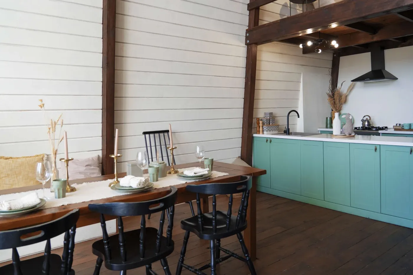 A rustic kitchen and dining area with a wooden table set for four, black chairs, green cabinets, and white shiplap walls, conveying a cozy, farmhouse feel.