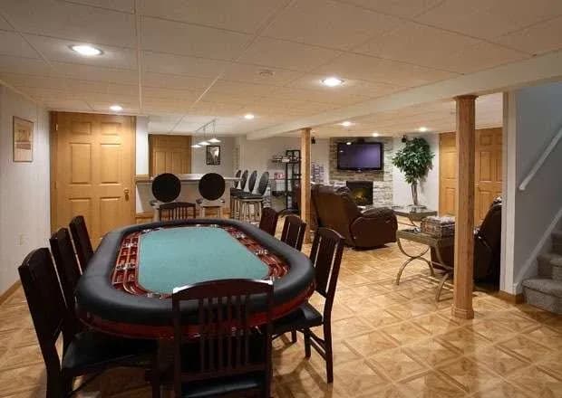 A cozy basement game room features a poker table with wooden chairs, a bar area with stools, a TV on the wall, and comfortable seating near a fireplace.