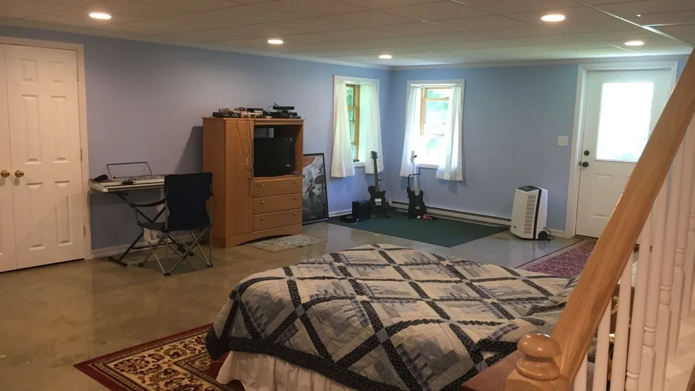 A spacious basement room with blue walls features a quilt-covered bed, a desk with a foldable chair, a wooden dresser with a TV, and musical instruments by the window.